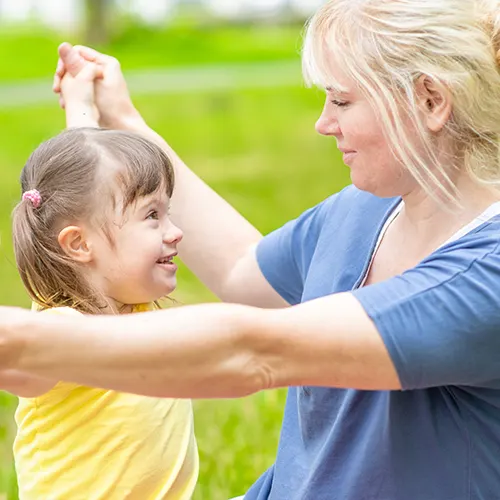 A smiling child with Down syndrome playing joyfully with a caregiver in a park, representing supportive and inclusive care.