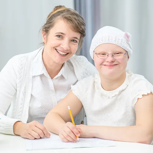 A young girl with Down syndrome smiling while writing with support from a caregiver, symbolising personalised NDIS continence and toilet training support.