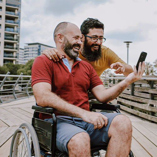 Two happy men outdoors, one sitting in a wheelchair and the other standing beside him, smiling and looking at a smartphone together — representing friendship, independence and NDIS community support in Sunshine, Melbourne.