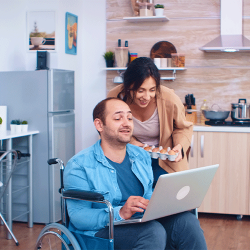 Smiling woman assisting a man in a wheelchair using a laptop at home, representing NDIS continence assessment and independent living support in Darwin, Northern Territory.