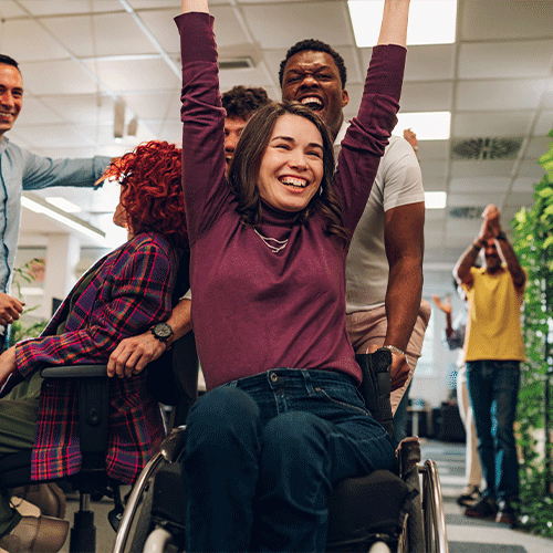 Group of colleagues celebrating as a woman in a wheelchair raises her arms and smiles in an inclusive workplace setting.