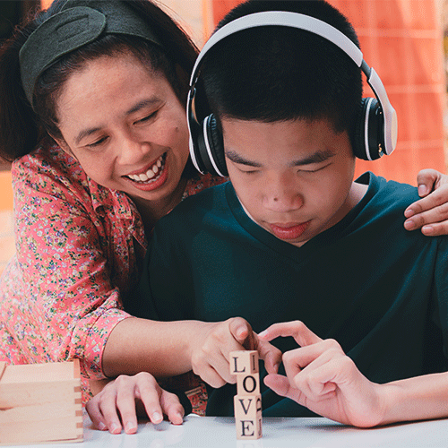 A support worker helping a child with headphones stack wooden blocks during an outdoor learning activity.