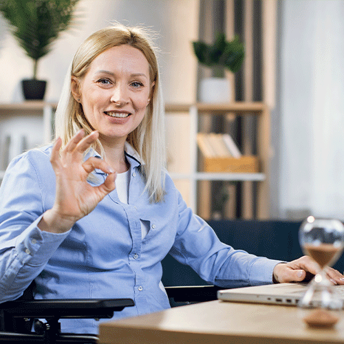 Woman in a wheelchair smiling and making an OK gesture while working on a laptop, representing continence assessment and disability support in Westcourt, Queensland.