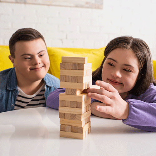 Two young people with Down syndrome smiling and playing a wooden block stacking game together at home – NDIS disability support services on the Central Coast
