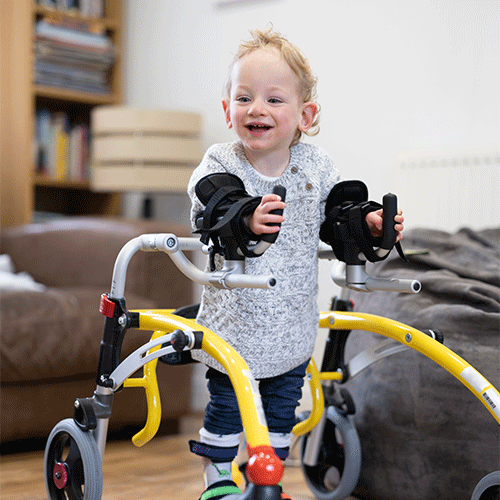 A smiling young child using a yellow paediatric gait trainer at home, supported by NDIS disability services