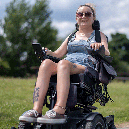 Young woman with disability smiling outdoors in a power wheelchair on a sunny day.