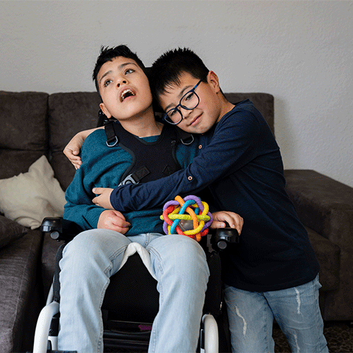 A young boy in a wheelchair with a chest support harness is hugged by his older brother on the couch, both smiling, for A1 Care Group continence and disability support services